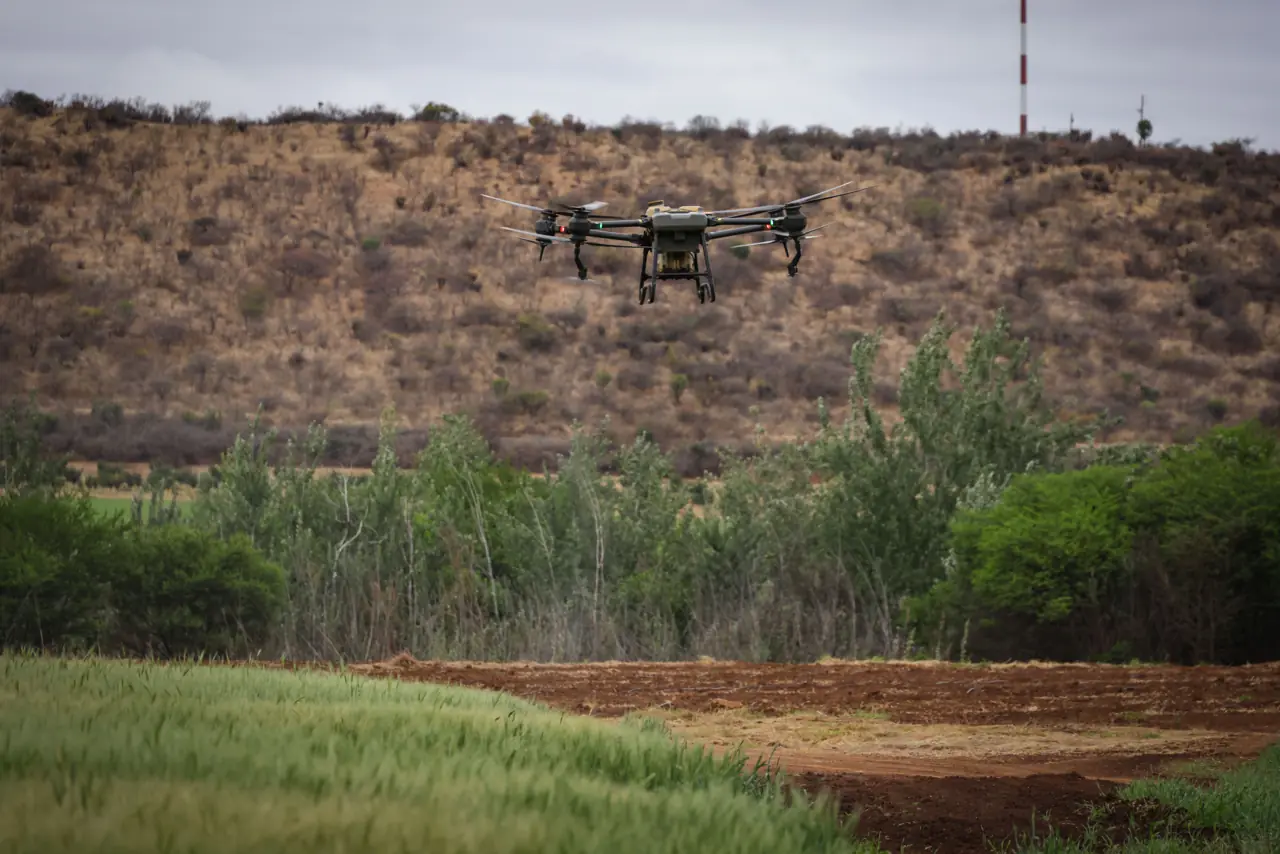 An agricultural drone in the air over a green field, hills behind